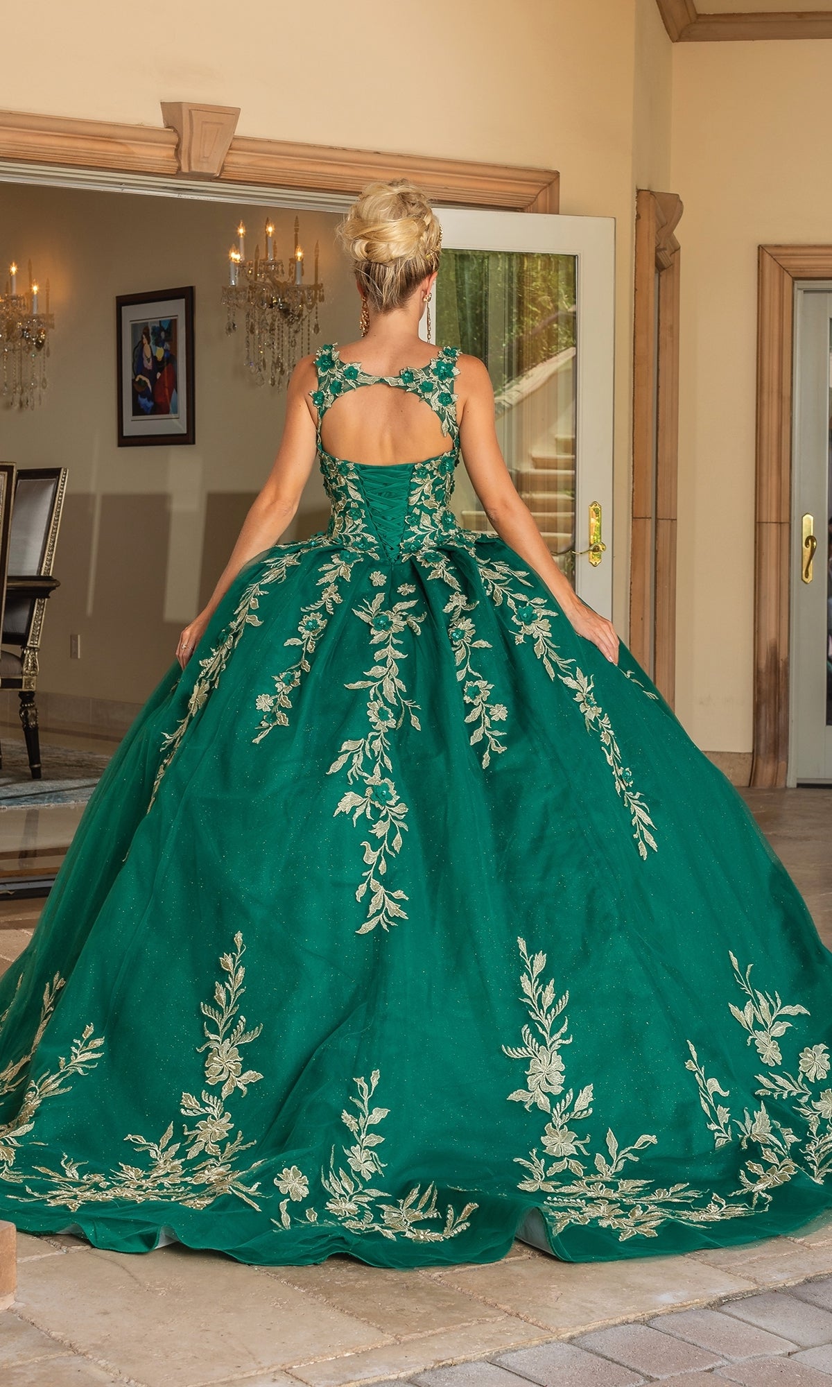 A woman with blonde hair in an updo wears the Quinceanera Dress 1755 By Dancing Queen in hunter green with gold floral embroidery and a sweetheart neckline, standing with her back to the camera in a refined indoor setting.
